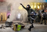 A riot police officer walks next to a burning street barricade flanked by a line of their colleagues.