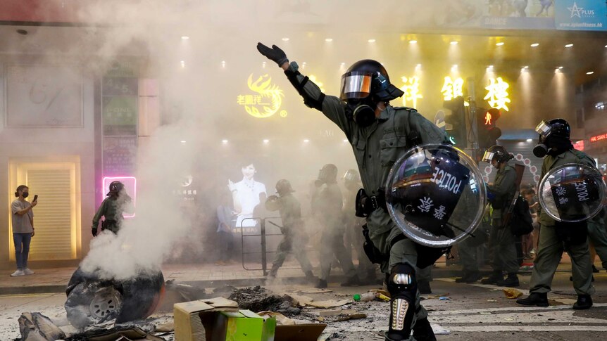 A riot police officer walks next to a burning street barricade flanked by a line of their colleagues.