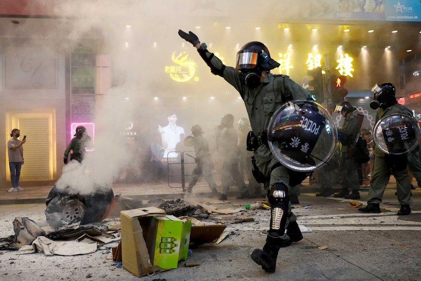 A riot police officer walks next to a burning street barricade flanked by a line of their colleagues.