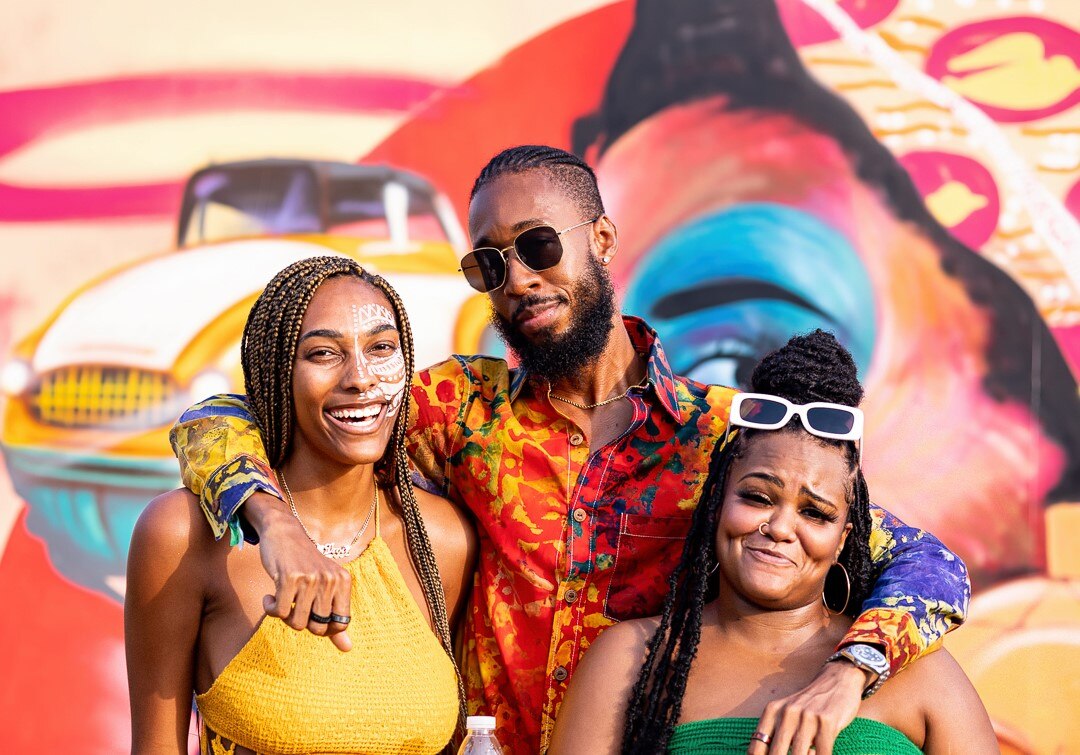 Three black people, a man and two women, arms around each other, wearing bright colours, happy