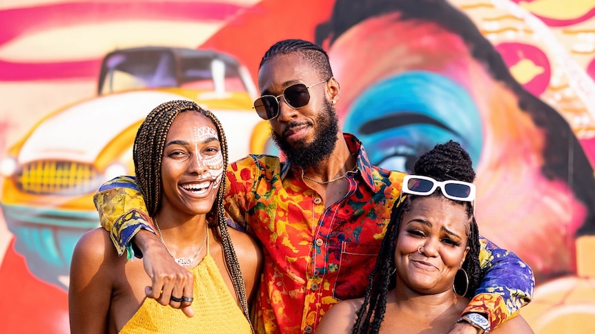 Three black people, a man and two women, arms around each other, wearing bright colours, happy
