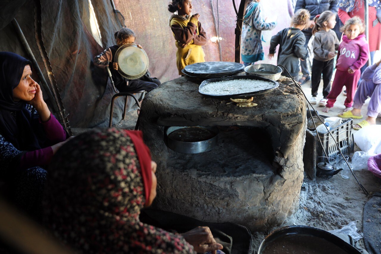 Duas mulheres estão sentadas perto de um forno de pedra e várias crianças podem ser vistas ao longe.
