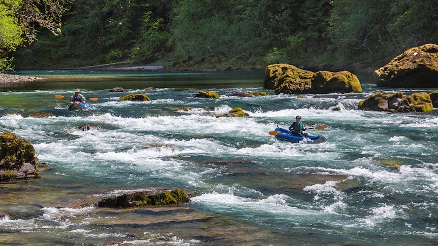 A landscape photograph of a vivid turbulent river with two canoeists navigating.