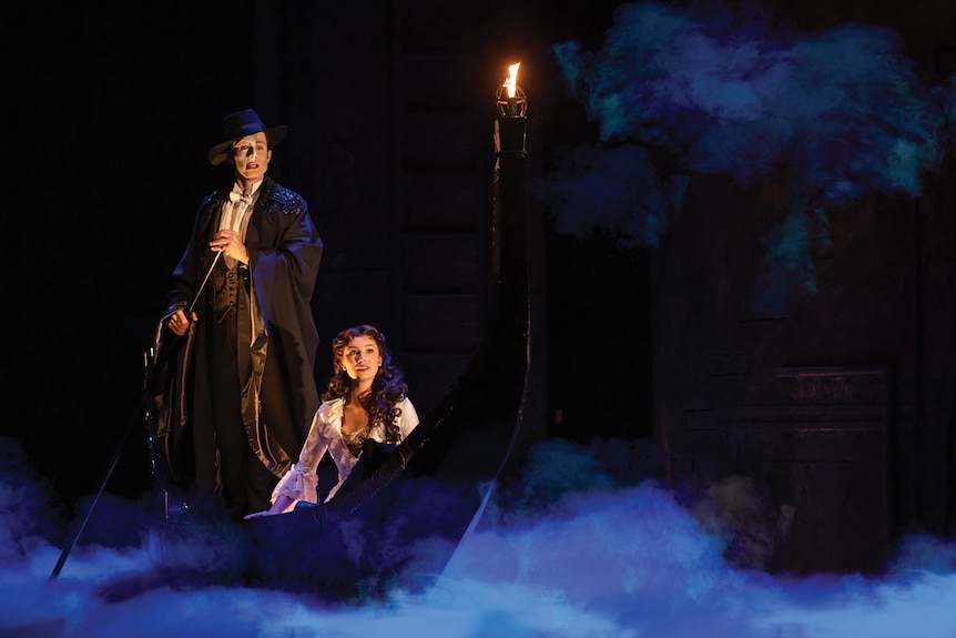 A white man in a mask and cape rows a boat with a white woman with dark hair across a smoke-filled stage.