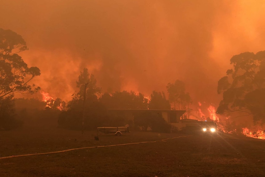 Firefighters work to save a property surrounded by smoke and with flames behind it.