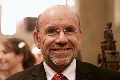 A balding man dressed in a black suit and red time smiles at the camera.