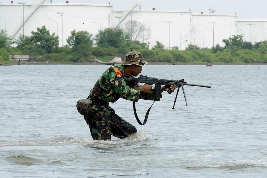 An Indonesian man in army gear stands pointing a gun in a body of water. 