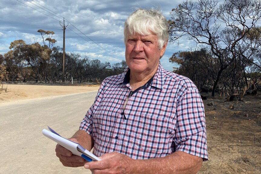 Older man in check shirt holding notebook, standing on edge of road in front of a power pole