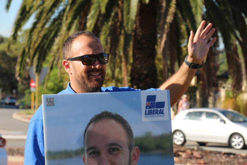 Liberal candidate Scott Edwardes wearing a blue t-shirt and sunglasses holds an election poster.