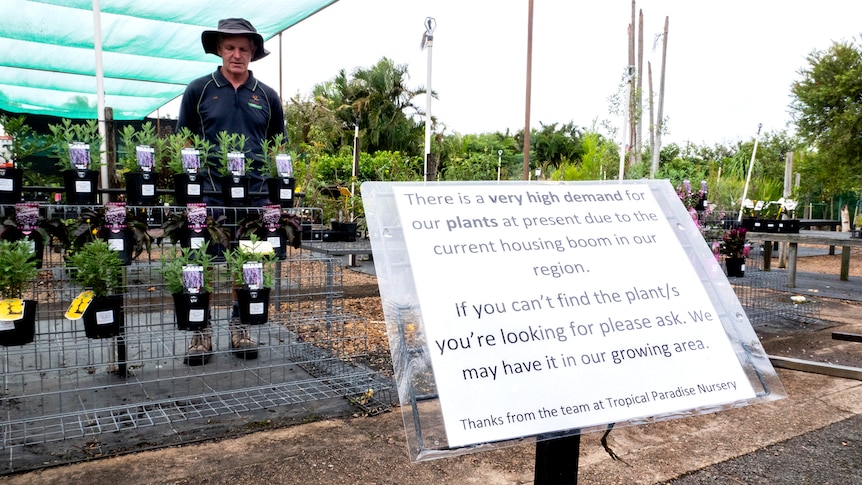 A man stands in a garden with a sign saying due to a housing boom and high demand they are short of plants.