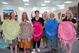 seven women standing in a line with colourful weaves in their hands 