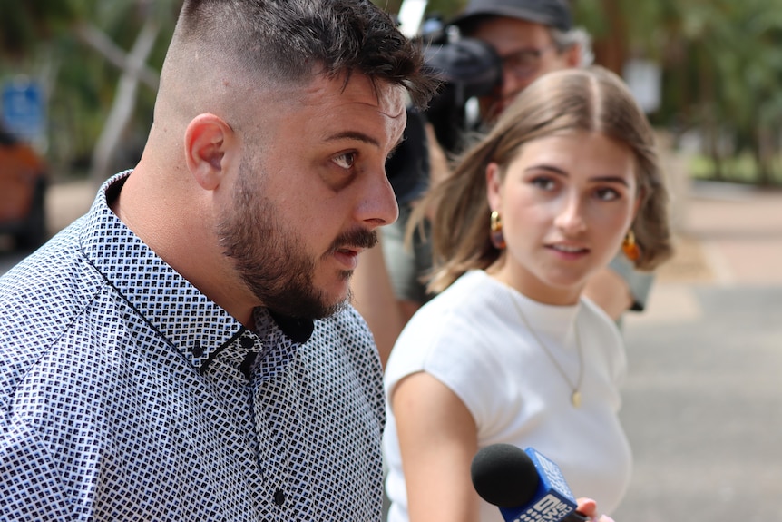 A man in a dress shirt walks as a woman holds a microphone under his face. 