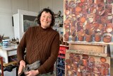 Artist Lucy Culliton smiles at the camera with artwork next to her, wearing a brown jumper