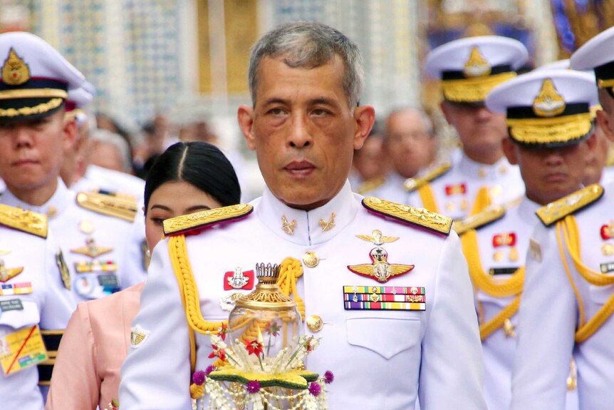 Thai King Maha Vajiralongkorn wears a decorative white and gold uniform and is walking in front of a large group of officials