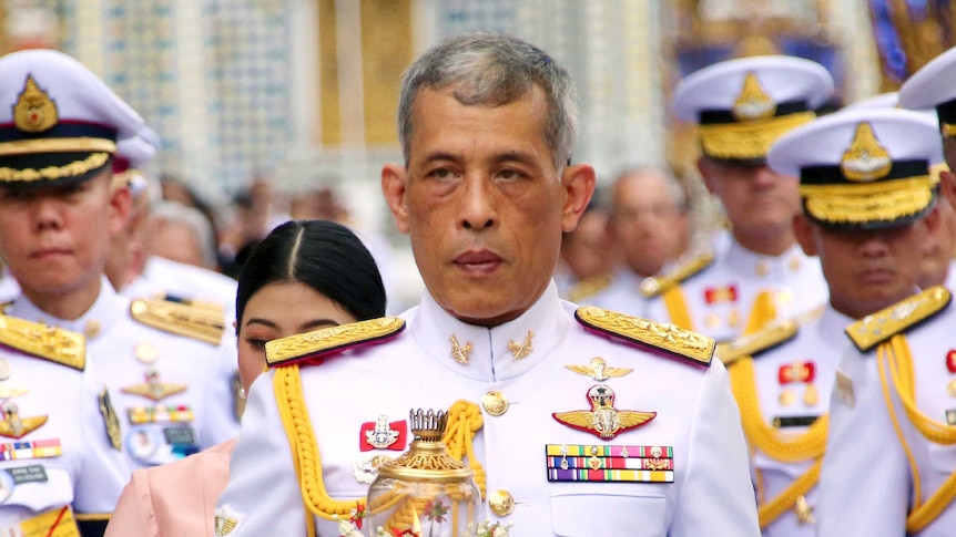 Thai King Maha Vajiralongkorn wears a decorative white and gold uniform and is walking in front of a large group of officials