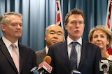 A group of poiticians stand at a lectern in front of Australian flags.