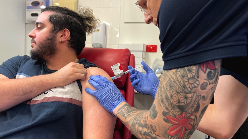 A nurse with a tattooed arm injects a bearded man in a hospital clinic