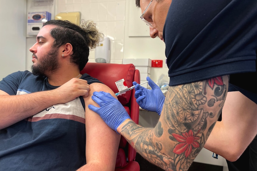 A nurse with a tattooed arm injects a bearded man in a hospital clinic