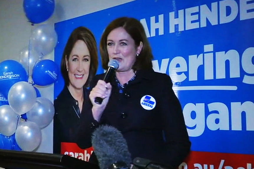 Sarah Henderson holds a microphone as she addresses a gathering in front of a poster and balloons.