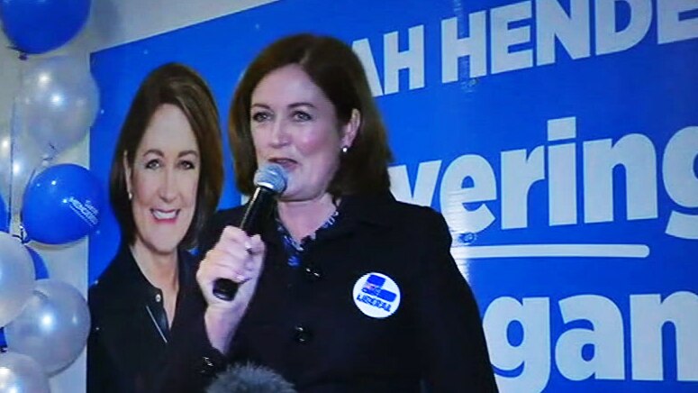 Sarah Henderson holds a microphone as she addresses a gathering in front of a poster and balloons.