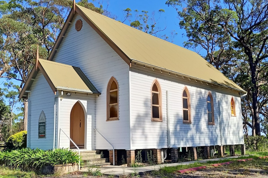 A white weatherboard church with beige roof, salmon-coloured windows, with blue sky and bush background.