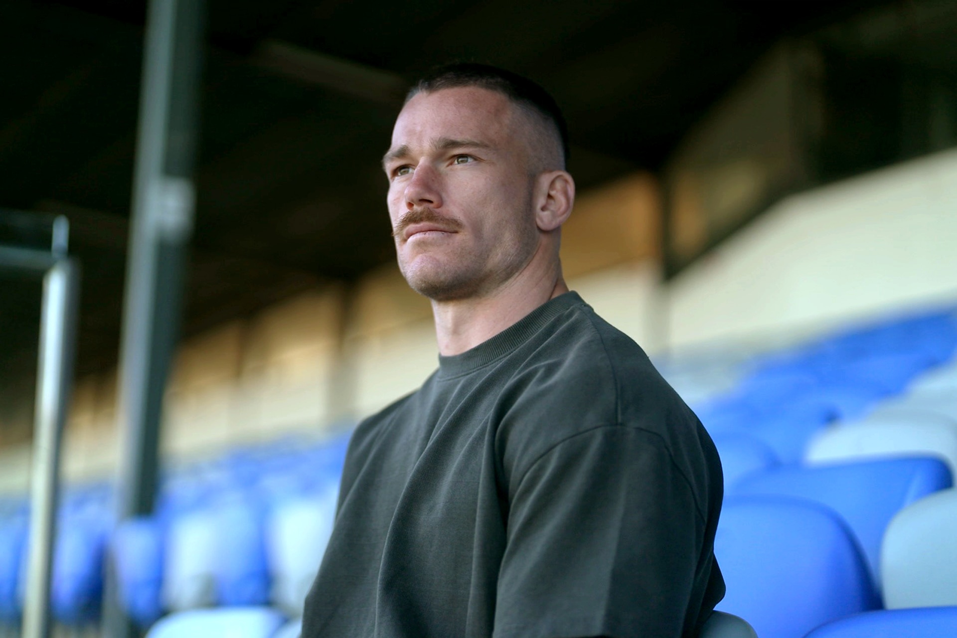 A man wearing a tshirt sits in a grandstand looking ahead with a determined expression.