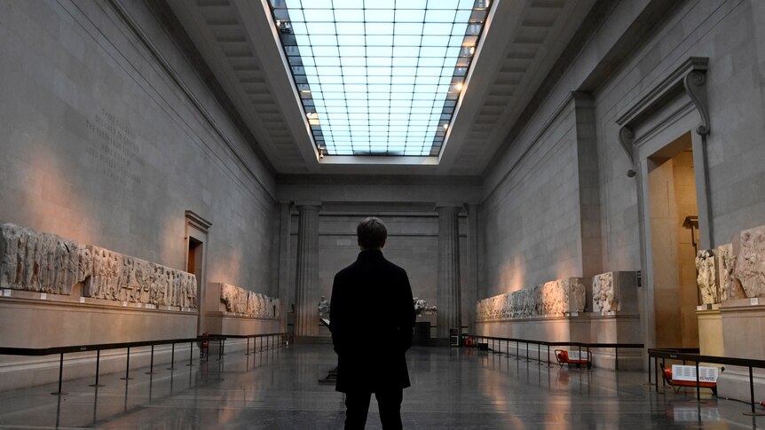 A man standing in the middle of a giant museum room with a skylight on the ceiling and artefacts all around the walls