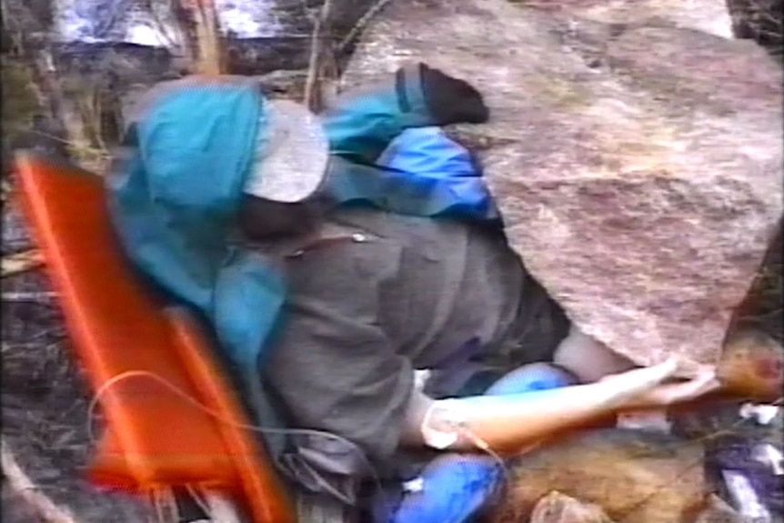 A man stuck underneath a boulder with a hat on his head.