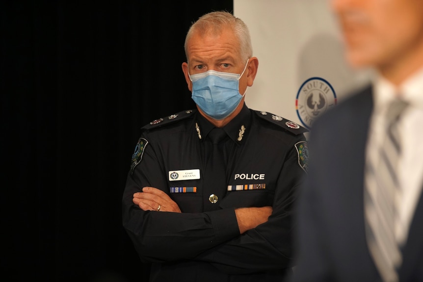 A man in police uniform and a blue surgical face mask stands with his arms folded