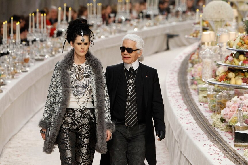 Stella Tennant is beside Karl Lagerfield walking past tables with candles and food on them. They have straight faces.