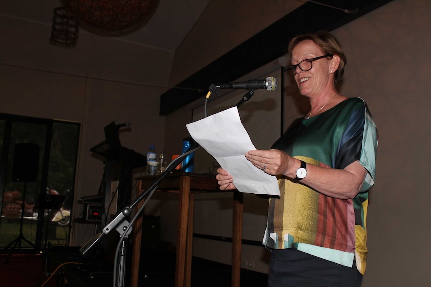 The Indigo mayor, Jenny O'Connor, stands on a stage and addresses a town forum on the water industry, holding a piece of paper.