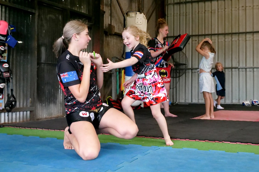 A young girl is pictured mid-movement in a muay thai class, an older teenager kneels next to her. 