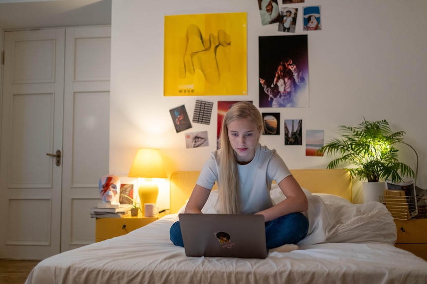 A teenage girl sits on her bed in front of a laptop.