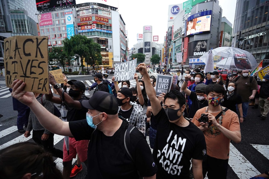 About 100 people are seen walking through a Tokyo street with buildings with advertisements on them behind the group.