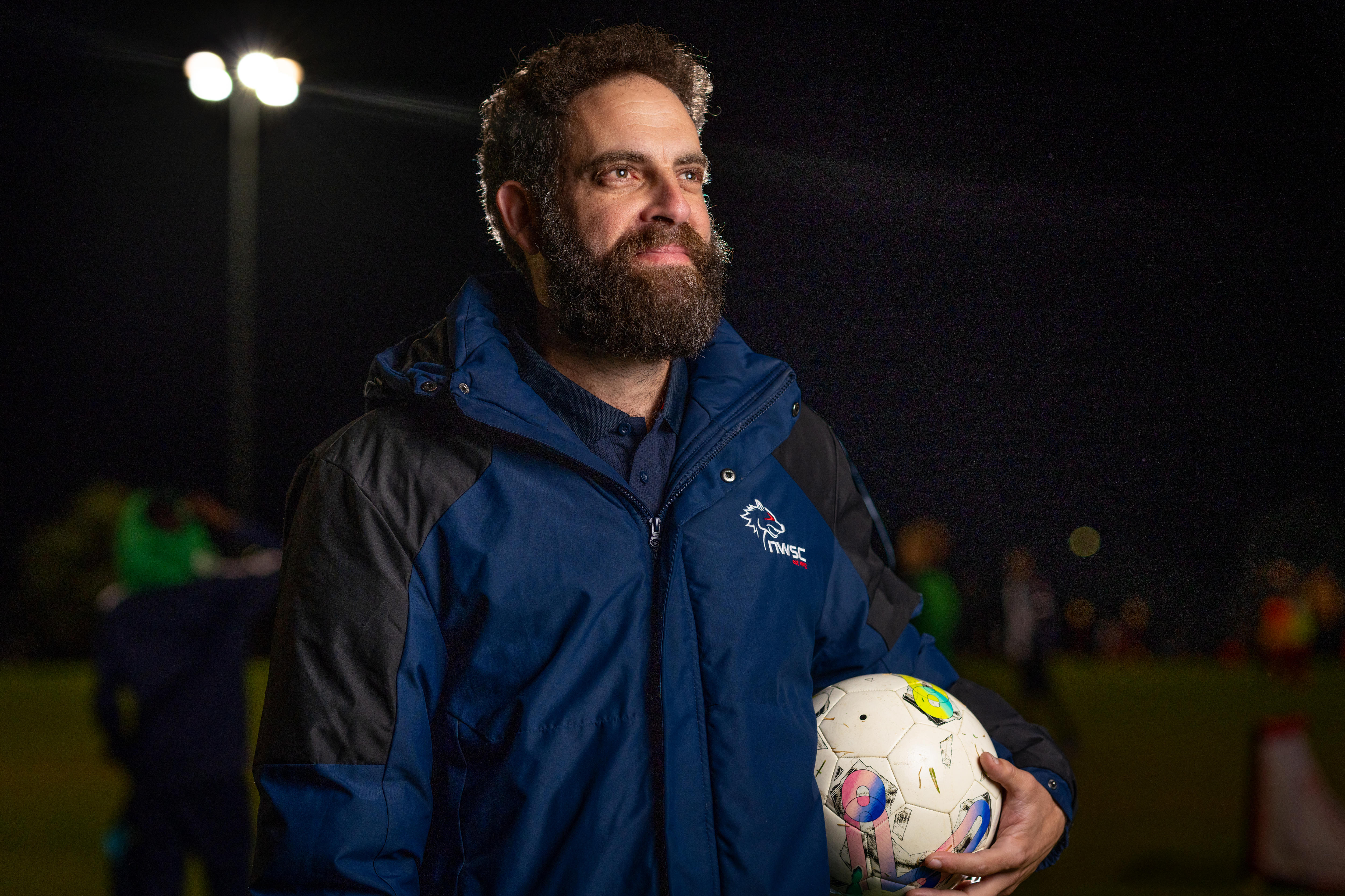 A man in a blue jacket holds a soccer ball at his waist.
