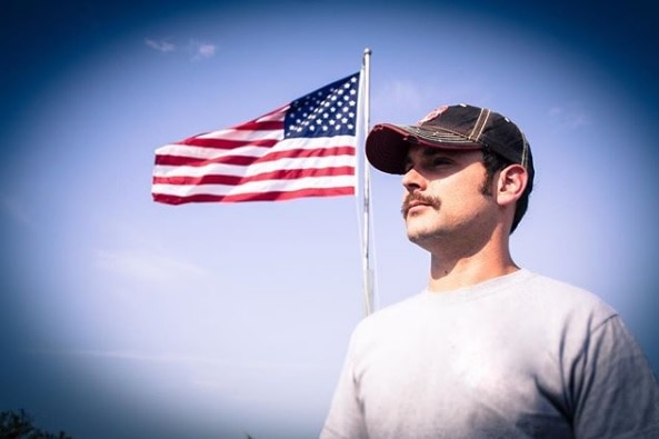 Josh Abbott Band guitarist Caleb Keeter standing in front of US flag with blue sky in the background