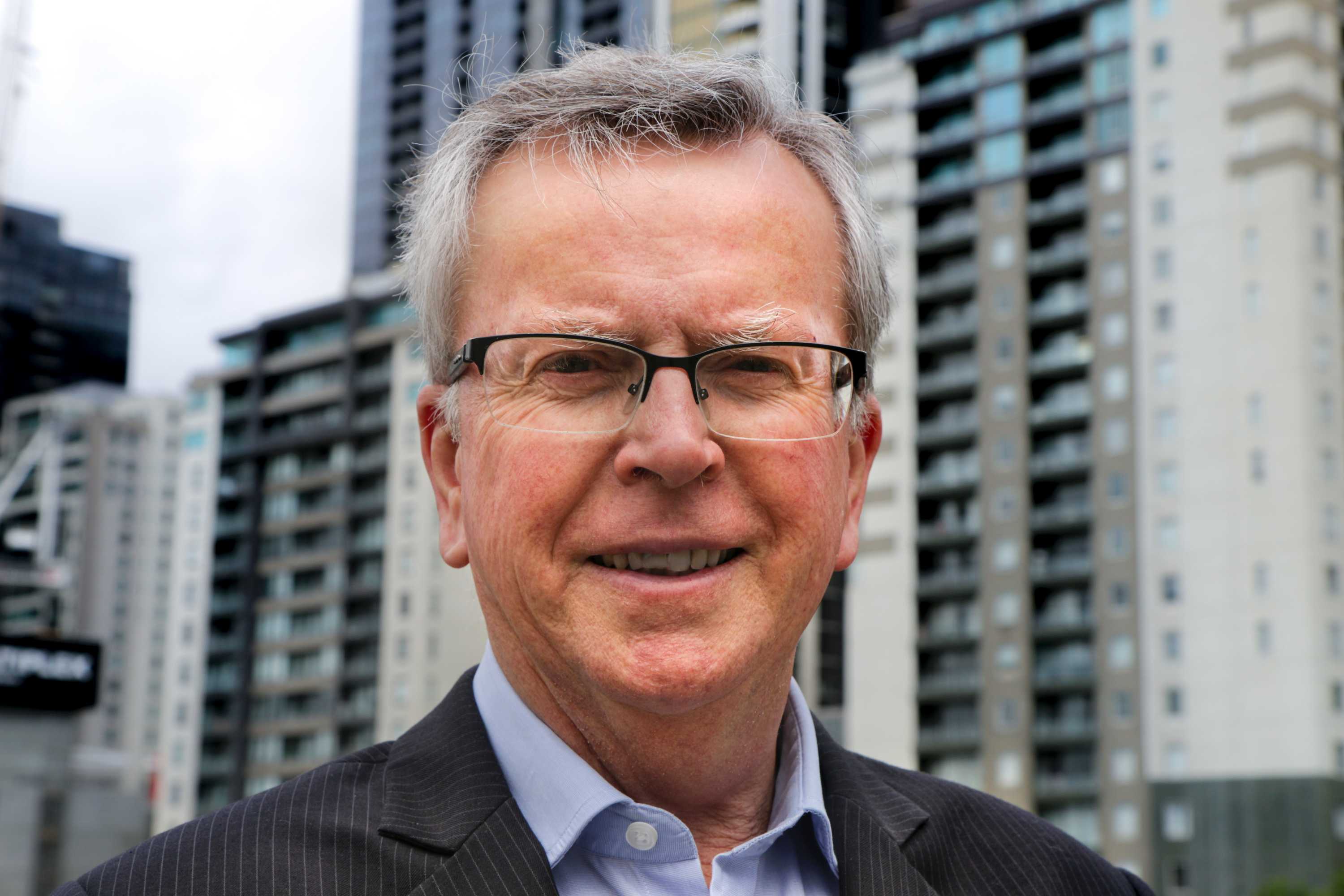 Man with glasses and grey hair stands outside in front of city buildings.