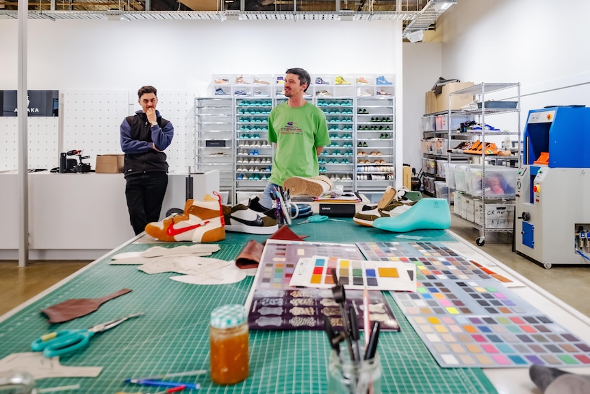 Chase Shiel stands in his workshop with an assitant and shoemaking materials around.