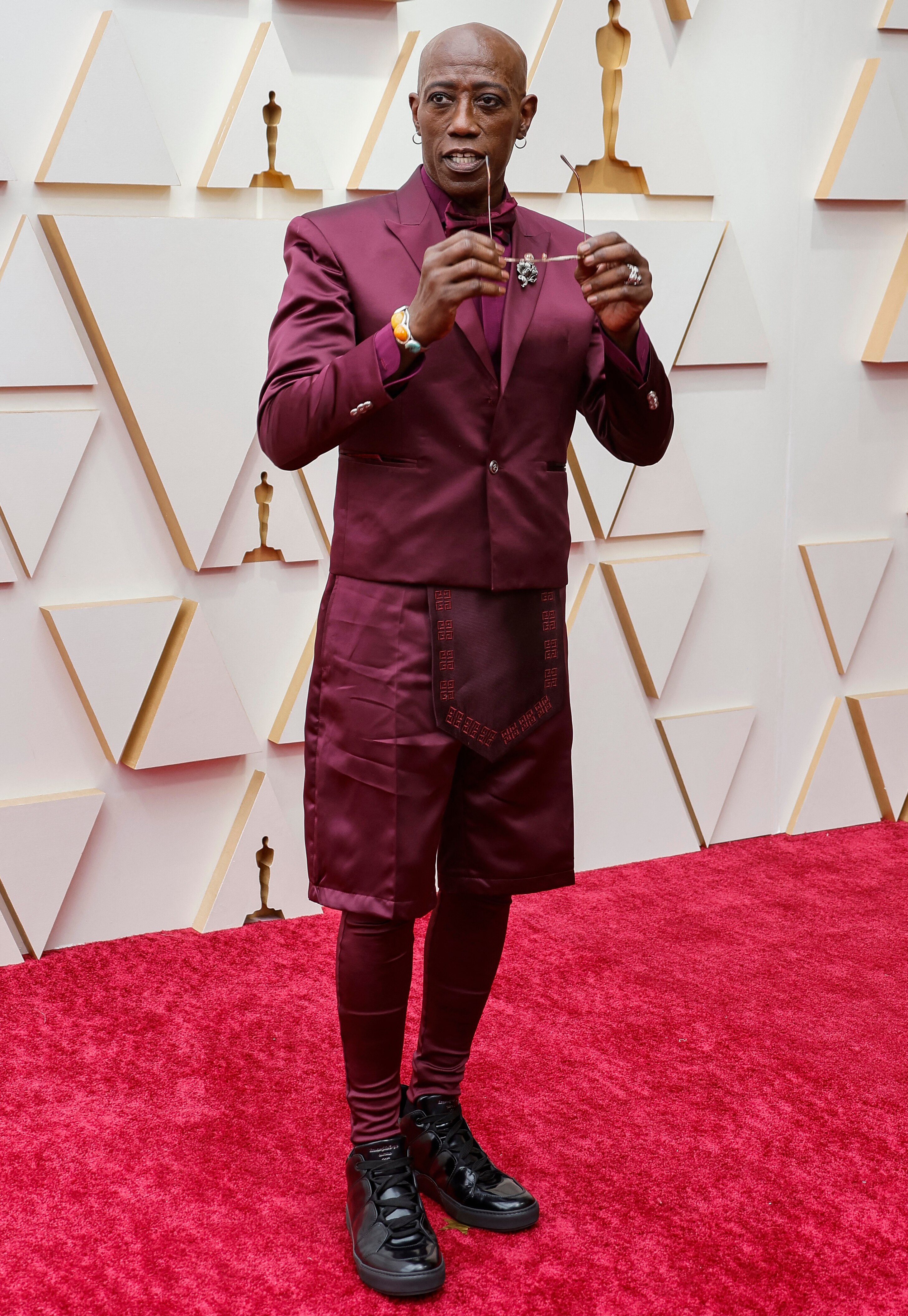 Actor Wesley Snipes poses on a red carpet holding a pair of sunglasses near his face, he is wearing a maroon ensemble and boots