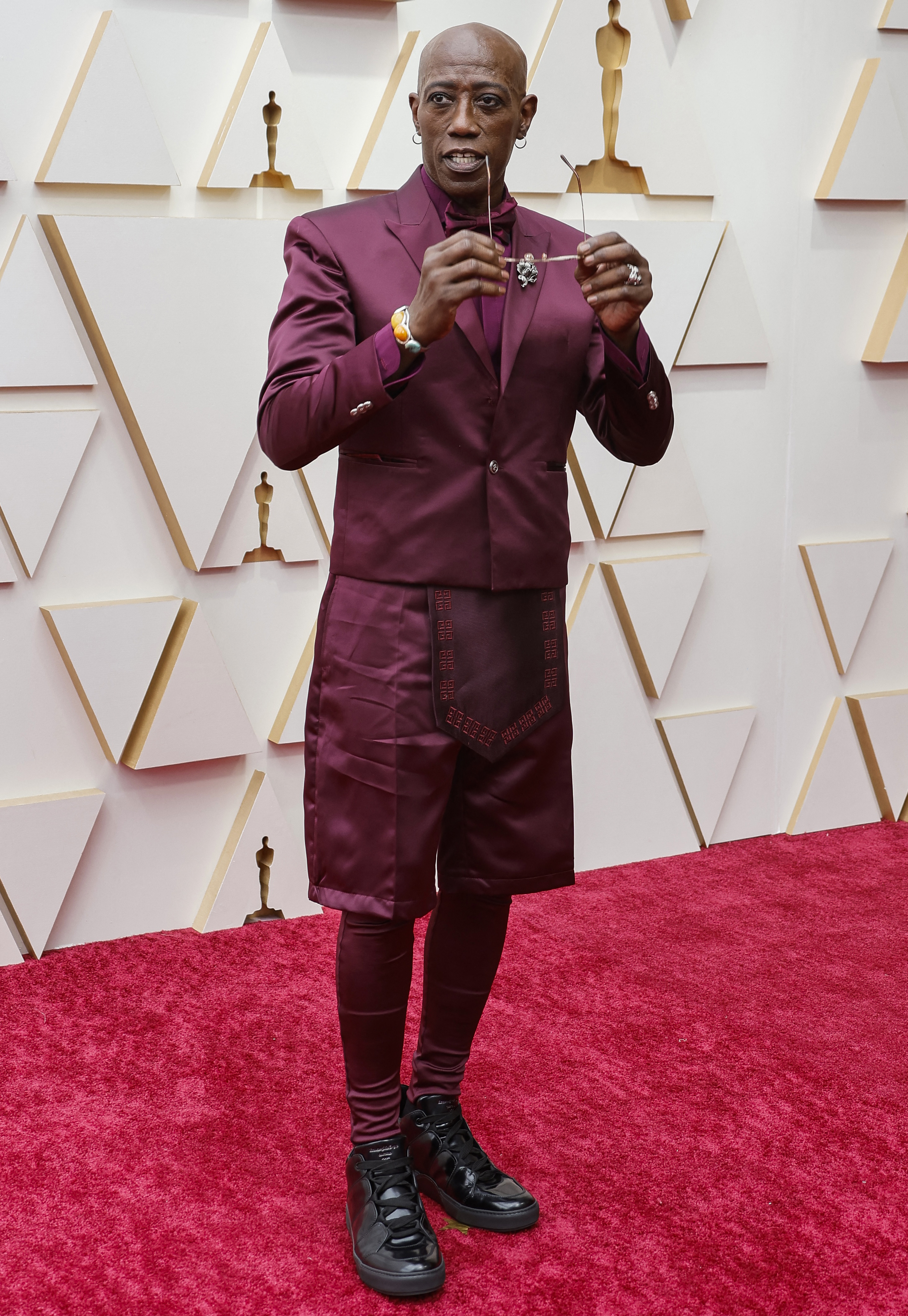 Actor Wesley Snipes poses on a red carpet holding a pair of sunglasses near his face, he is wearing a maroon ensemble and boots
