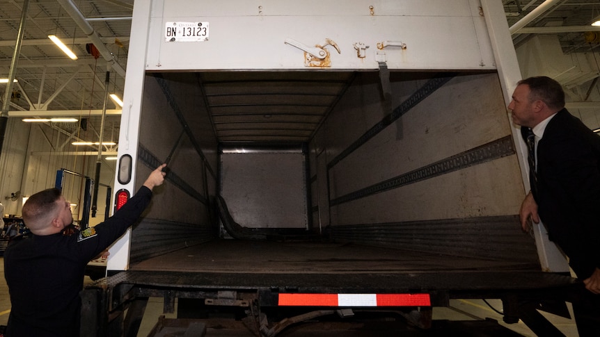 Two people stand behind a large truck and one points at the inside which is empty