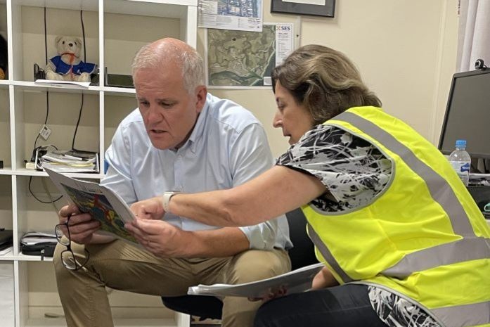 Prime Minister Scott Morrison wears work boots and looks at a document. A woman wearing high-vis looks on.