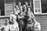 A black and white photo of a group of soldiers in uniform smiling in front of a weatherboard house.