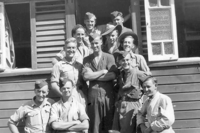 A black and white photo of a group of soldiers in uniform smiling in front of a weatherboard house.