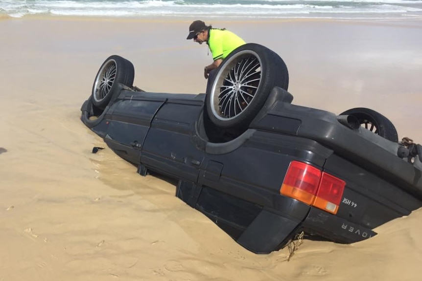 A man inspecting a car upside down and half-buried in the sand