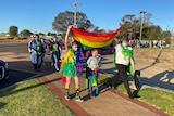 Three people, holding up a rainbow flag, walk along a path in front of a crowd of people. 