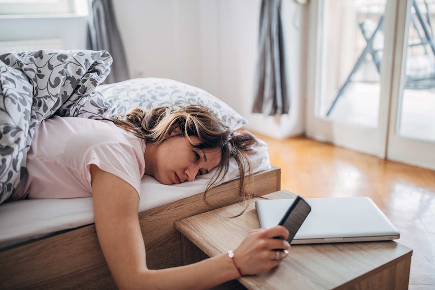 A woman in bed looks tiredly at her smart phone.