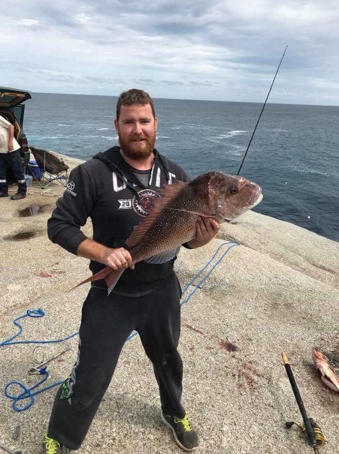 A man with a beard standing on rocks holding a fish, with ocean in the background.