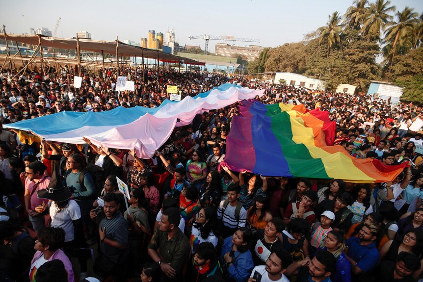 A crowd of people carry giant transgender and LGBT pride flags over their heads. 