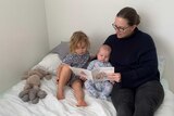 Jessie reads a book to her two children while sitting on a bed.